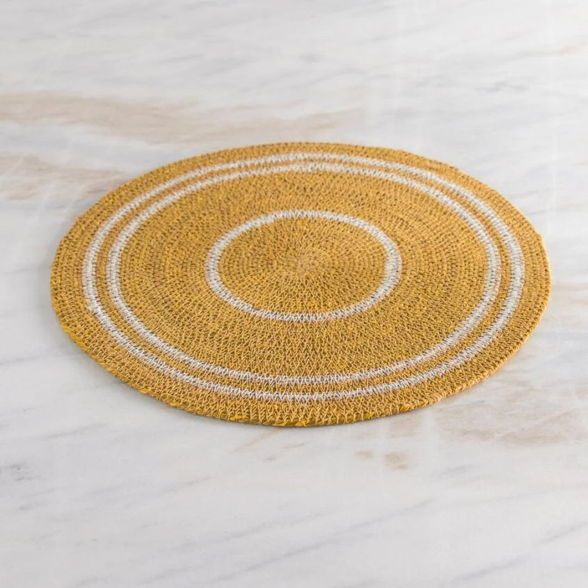 A round woven placemat in mustard yellow with concentric white rings, displayed on a light marble surface.