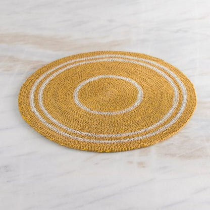 A round woven placemat in mustard yellow with concentric white rings, displayed on a light marble surface.