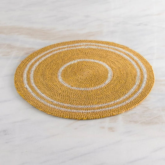 A round woven placemat in mustard yellow with concentric white rings, displayed on a light marble surface.