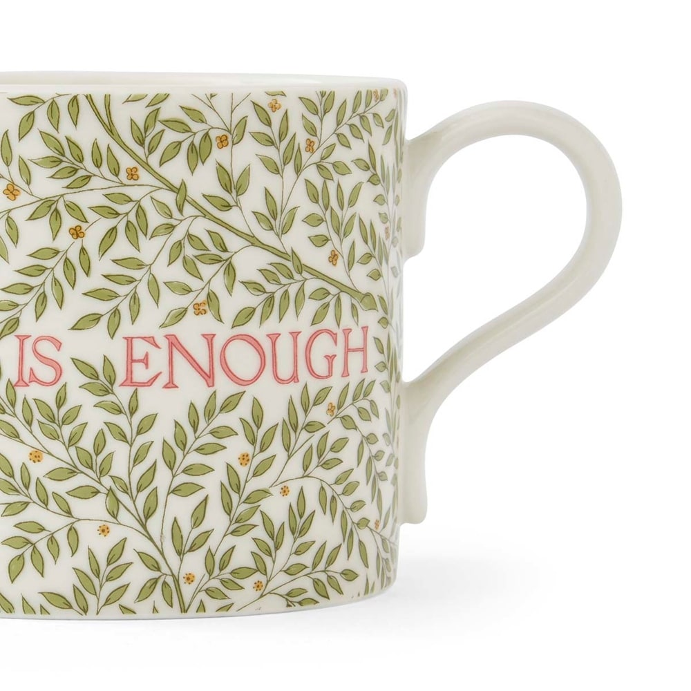 A close-up shot of a white ceramic mug with a traditional curved handle and featuring a Morris & Co. pattern of green leaves and small yellow flowers, with the words "IS ENOUGH" printed in bold red capital letters across the front.
