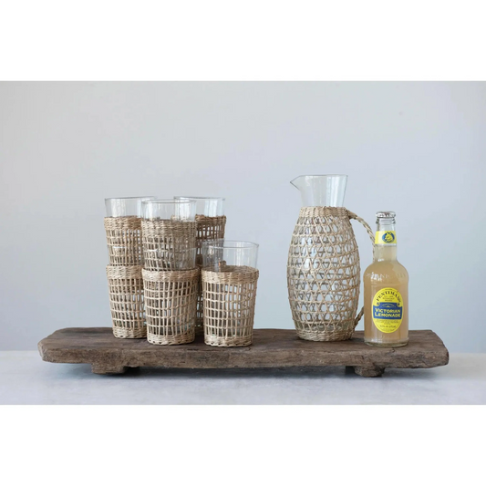 A few stacks of glasses with natural woven sleeves, a glass pitcher with a natural woven sleeve and a bottle of 'Victoria Lemonade' on a footed wood board.