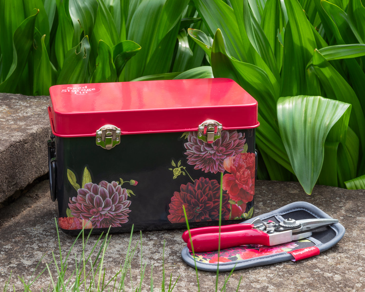 Floral-patterned storage tin and garden shears on a stone surface in front of green foliage.
