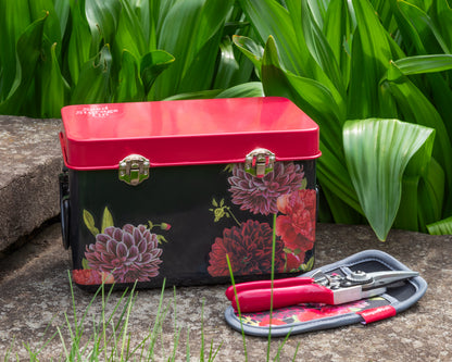 Floral-patterned storage tin and garden shears on a stone surface in front of green foliage.