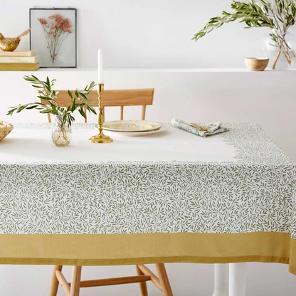 A styled dining table features a white tablecloth adorned with a dense, green leafy pattern along its border, finished with a solid yellow band at the hem. On top of the table sits a slender white candle in a brass holder, a small glass vase with green foliage, a stack of ceramic plates, and a matching patterned cloth napkin. In the background, a wooden chair rests against a light wall, next to a framed picture of dried red flowers and additional decorative items.