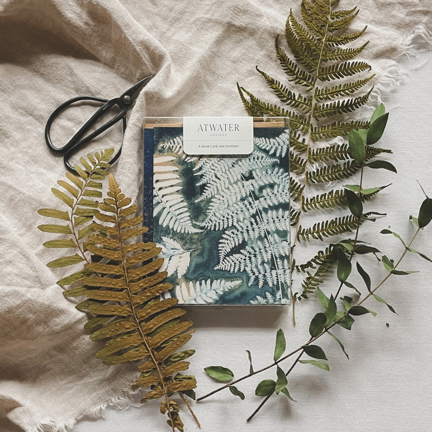 Fern cyanotype notecard set packaged in a clear box and surrounded by plants and garden sheers on a linen cloth